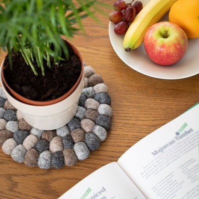 This handcrafted felted wool trivet combines a warm colour design with durable, sustainable materials.

Its hardwearing construction offers reliable protection for your surfaces while adding an elegant bright touch of colour to your home.  Shown with a  green leafed plant, fresh fruit on a wooden table. 