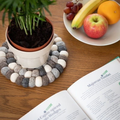 This handcrafted felted wool trivet combines a warm colour design with durable, sustainable materials.

Its hardwearing construction offers reliable protection for your surfaces while adding an elegant bright touch of colour to your home.

Shown with a glass pitcher jug of lemon water with a fresh salad and white bone china on a wooden table. 

Shown with a green leafed plant, a plate of fresh fruit with a cookery book on a wooden table. 
