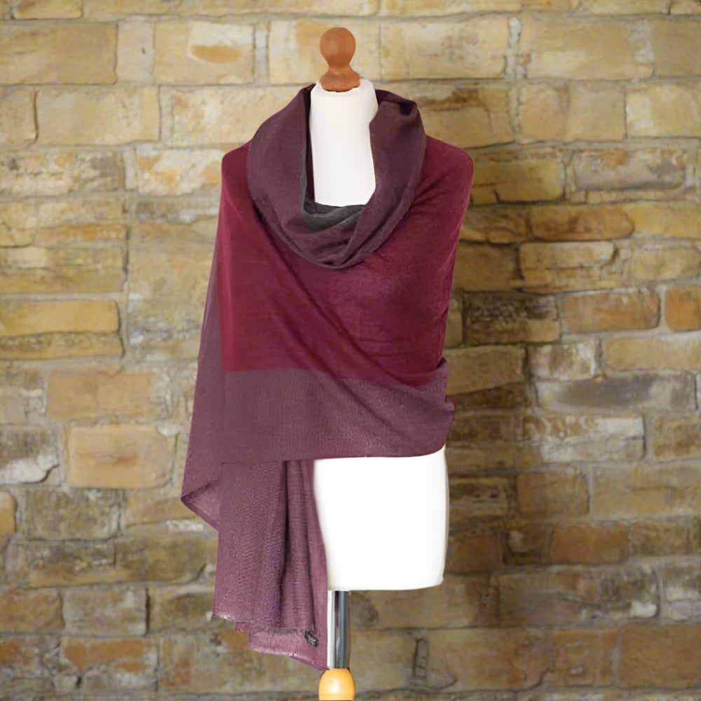 A Handwoven Cashmere Reversible Pashmina in Claret & Grey colours on a mannequin against a white background with a green plant decoration. 