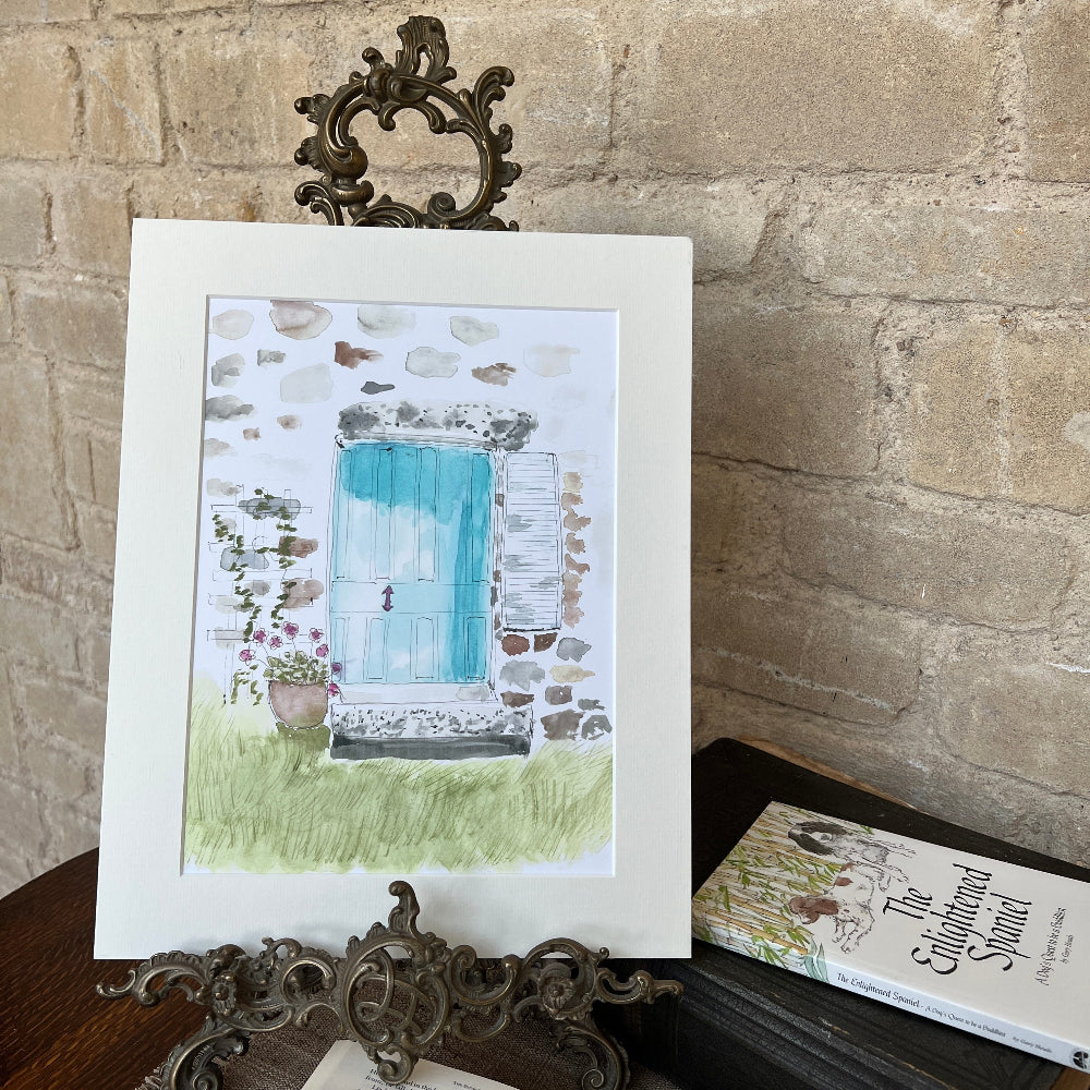Limited Edition Mounted Print &#39;A Cottage Doorway&#39; - by Linda Beazley shown on a brass stand, with a mug of coffee against a stone wall background with Limelight Candles room diffuser. 