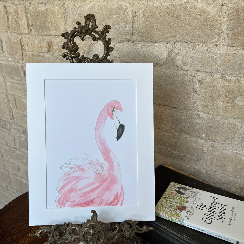 ‘Pink Flamingo’ is a limited edition watercolor print by Linda Beazley, elegantly presented on a mounted backing. This exquisite piece brings a touch of refinement to any environment and serves as an ideal gift or a treasured enhancement to your art collection.  Shown on a brass stand against a stone wall background with books, coffee and a Limelight Candles room diffuser. 