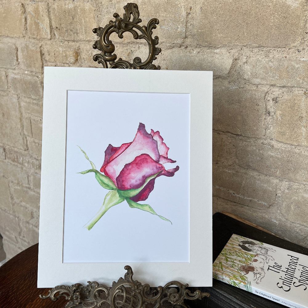 A Rose for My Love – Limited Edition Watercolour Print by Linda Beazley shown on a brass stand, against a stone wall background and a copy of the Enlightened Spaniel book.