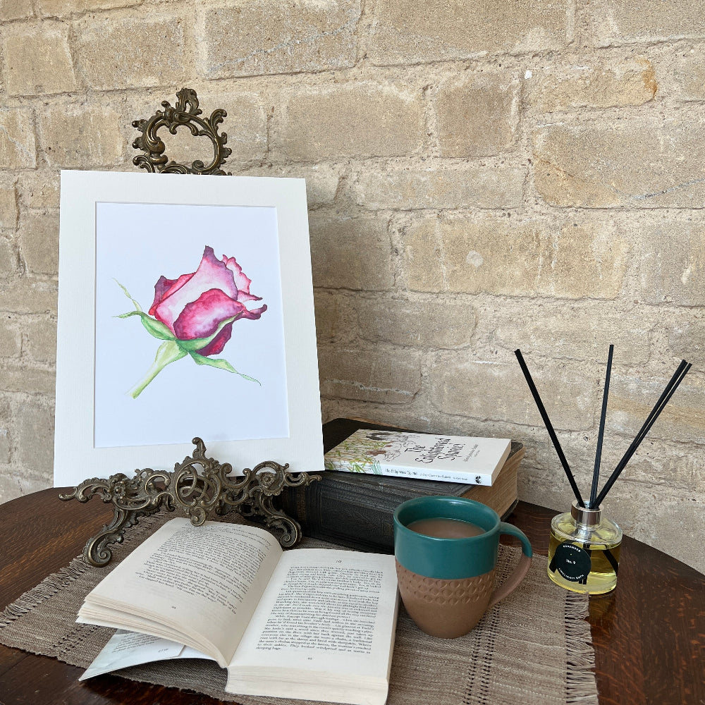 A Rose for My Love – Limited Edition Watercolour Print by Linda Beazley shown on a brass stand, against a stone wall background and a copy of the Enlightened Spaniel book.