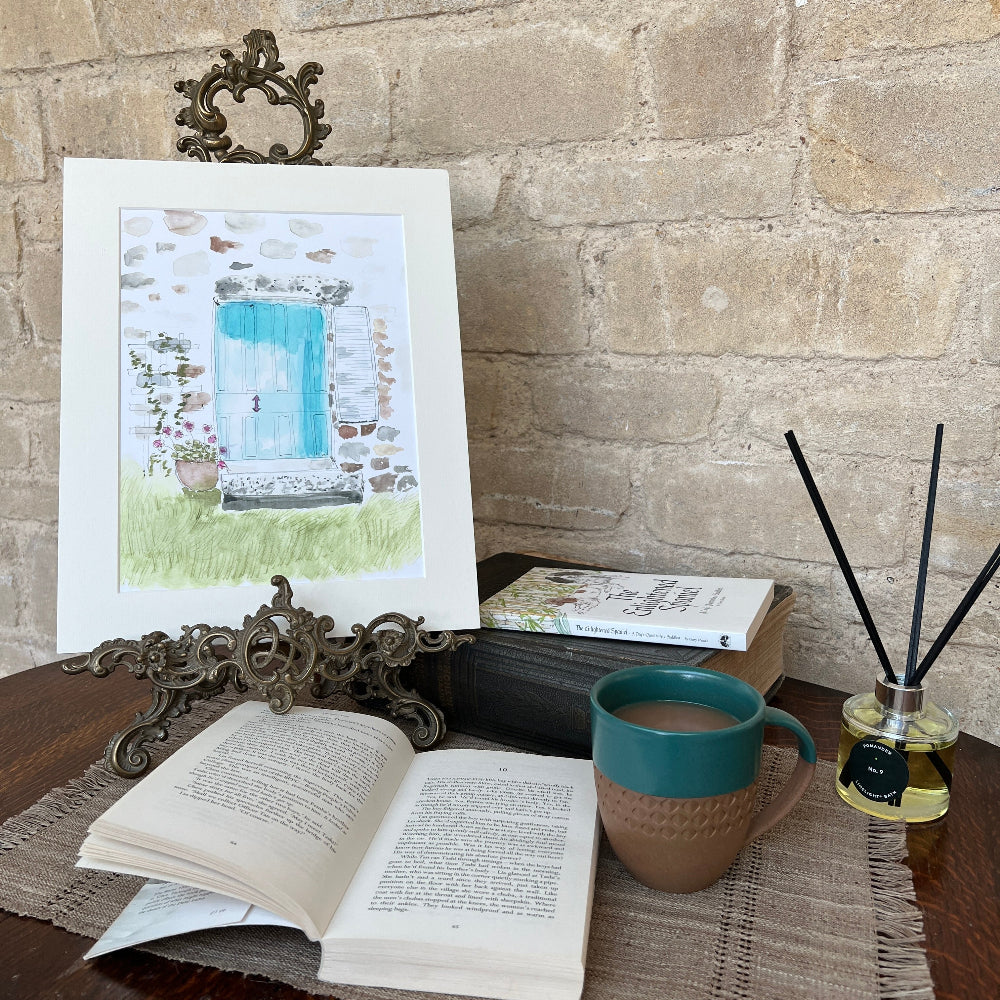 Limited Edition Mounted Print 'A Cottage Doorway' - by Linda Beazley shown on a brass stand, with a mug of coffee against a stone wall background with Limelight Candles room diffuser. 