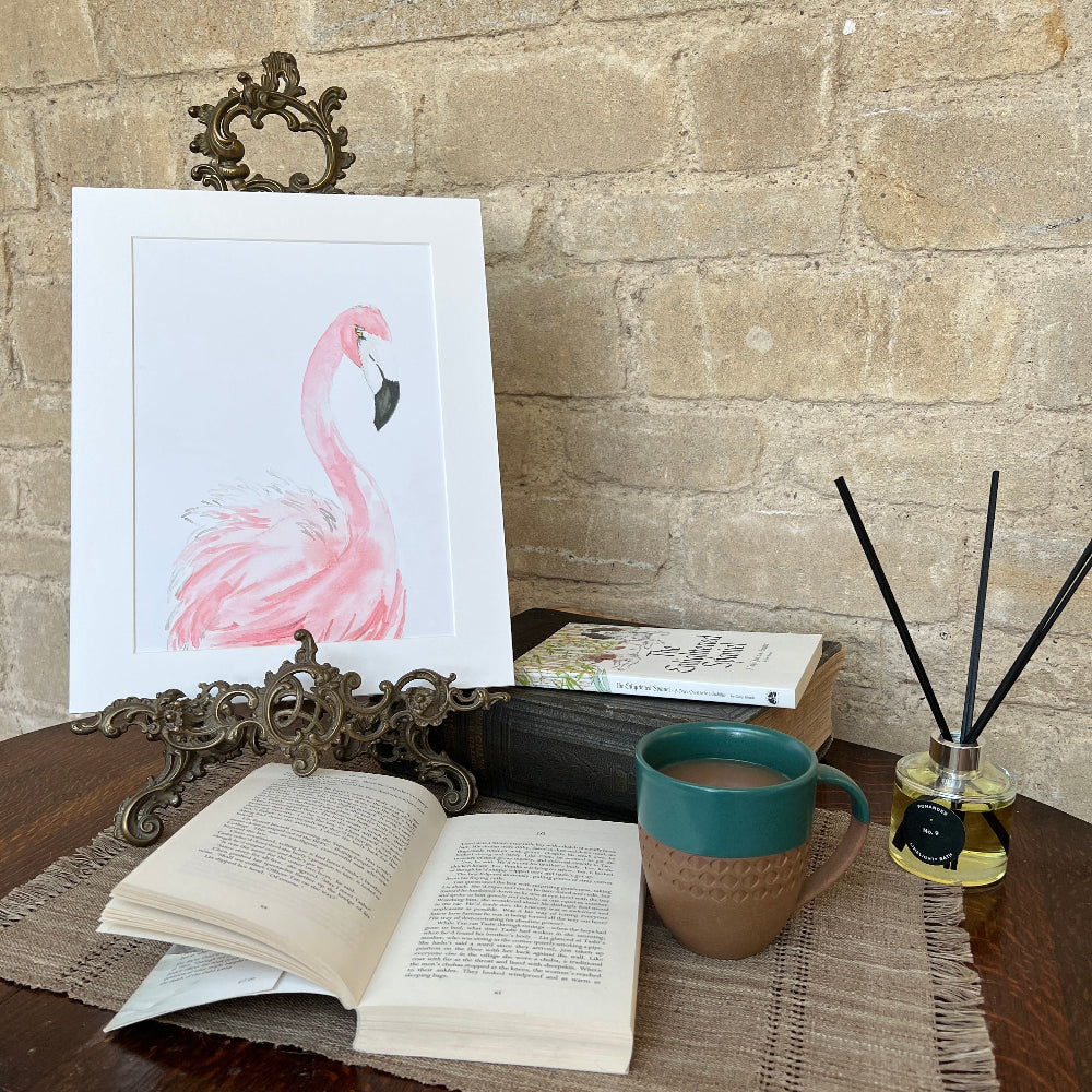 ‘Pink Flamingo’ is a limited edition watercolor print by Linda Beazley, elegantly presented on a mounted backing. This exquisite piece brings a touch of refinement to any environment and serves as an ideal gift or a treasured enhancement to your art collection.  Shown on a brass stand against a stone wall background with books, coffee and a Limelight Candles room diffuser. 