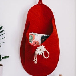 Pure Wool Felted Hanging Storage Bag with appliqued floral decoration in Red.  Shown with magazines against a white background with guitars in the room and a green potted plant.
