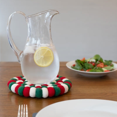This handcrafted felted wool trivet combines a strong festive red, white &amp;amp; green design with durable, sustainable materials.

Its hardwearing construction offers reliable protection for your surfaces while adding an elegant bright touch of colour to your home. Shown on a wooden surface with a glass pitcher of lemon water,  against a grey wall. 