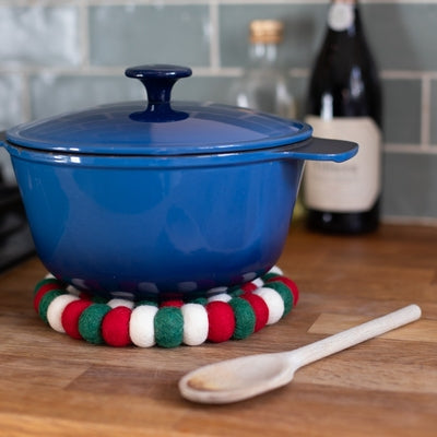 This handcrafted felted wool trivet combines a strong festive red, white &amp; green design with durable, sustainable materials.

Its hardwearing construction offers reliable protection for your surfaces while adding an elegant bright touch of colour to your home. Shown on a wooden surface with a stock pot, wooden spoon against a grey wall. 