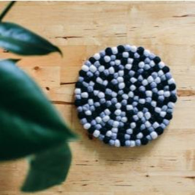 A round handcrafted sustainable wool trivet in black and grey colors, placed on a wooden surface with a green plant in the background.