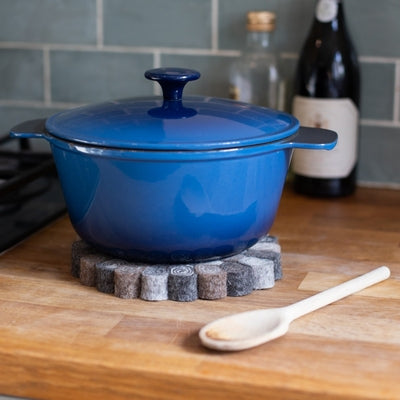 This handcrafted felted wool trivet combines a warm colour design with durable, sustainable materials. Shown with a blue casserole, a wooden spoon, olive oil against a grey tiled wall. 