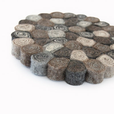 This handcrafted felted wool trivet combines a warm colour design with durable, sustainable materials. Shown with a blue casserole, a wooden spoon, olive oil against a grey tiled wall. Close up.