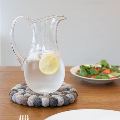 This handcrafted felted wool trivet combines a warm colour design with durable, sustainable materials.

Its hardwearing construction offers reliable protection for your surfaces while adding an elegant bright touch of colour to your home.

Shown with a glass pitcher of lemon water on a wooden table with fresh salad and white bone china. 