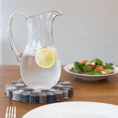 This handcrafted felted wool trivet combines a warm colour design with durable, sustainable materials. Shown with a glass pitcher jig with lemon water, fresh salad, white bone china on a wooden table. 